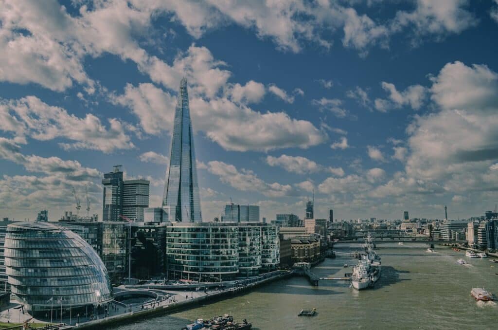 Horizonte da cidade com edifícios altos e modernos, The Shard, que é um arranha-céu de vidro, e um rio com barcos sob um céu parcialmente nublado.