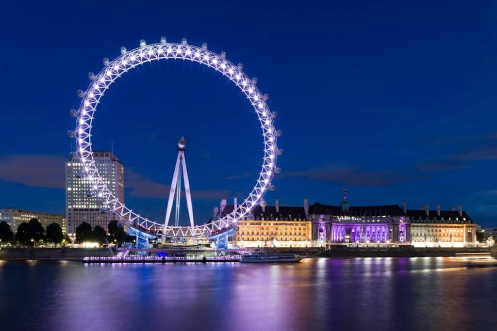 Roda gigante iluminada e edifícios perto de um rio à noite, com reflexos na água e um céu azul escuro e claro.