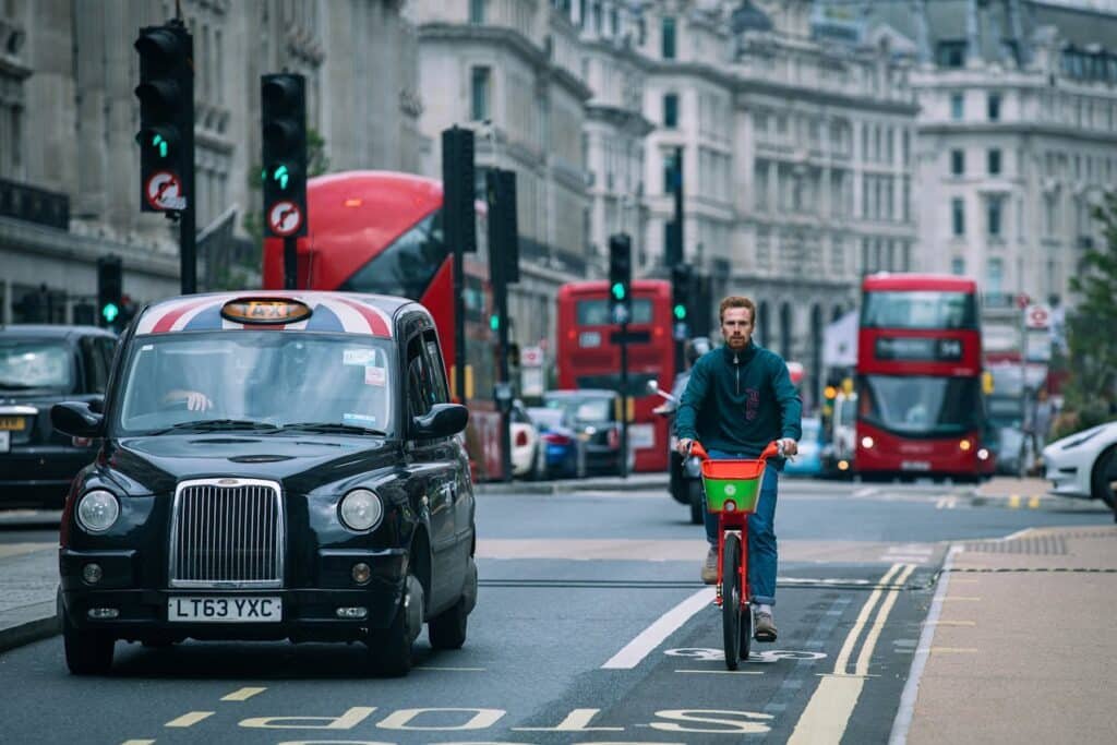 Um ciclista anda de bicicleta vermelha em uma rua de Londres com trânsito, incluindo um táxi preto e ônibus vermelhos de dois andares.
