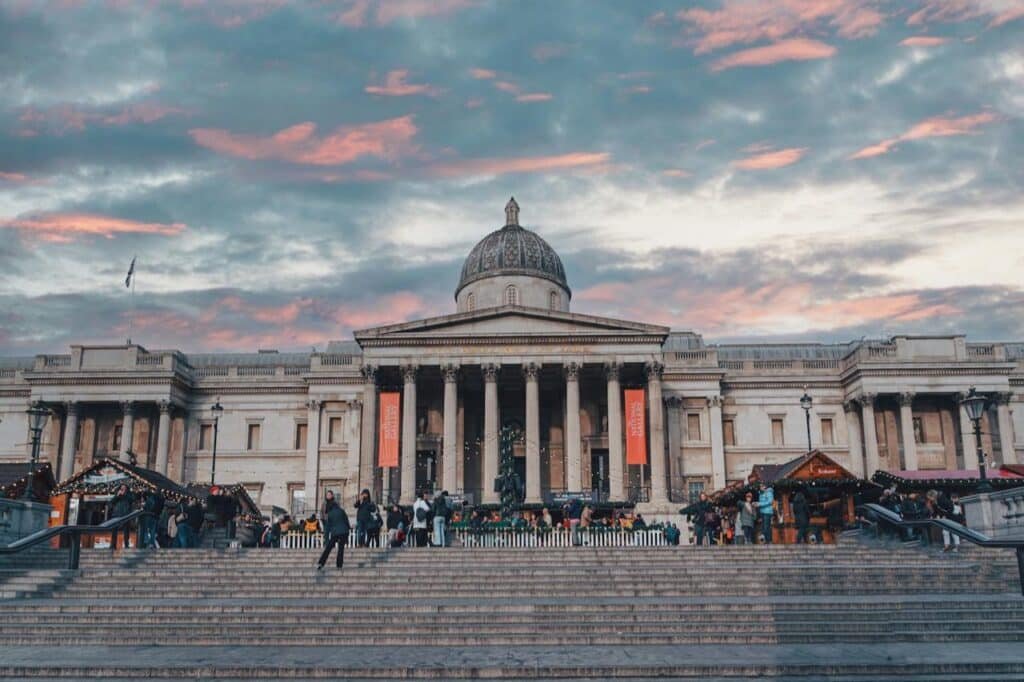A imagem mostra um edifício neoclássico com colunas e uma cúpula, pessoas nos degraus e um céu pôr do sol ao fundo. Essa é a National Gallery, uma das opções de o que fazer em Londres.