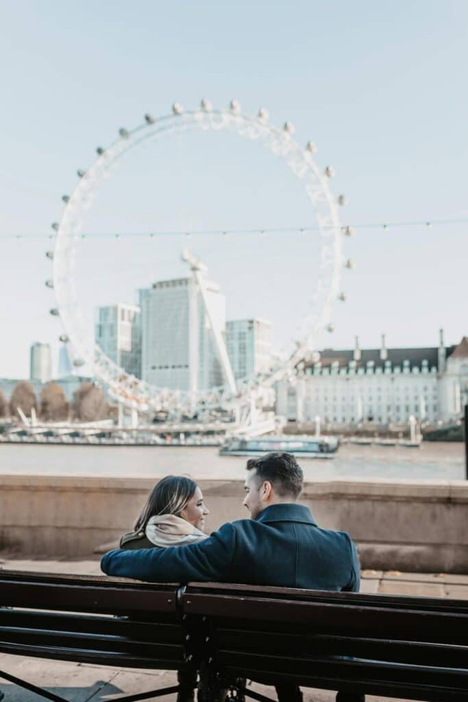 Um casal sentado em um banco perto do rio, com a London Eye e os prédios da cidade visíveis ao fundo.