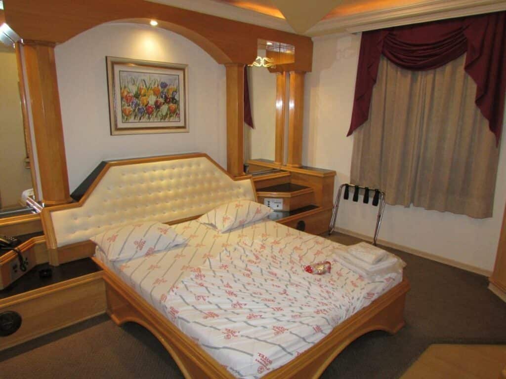 Quarto do Pleasant Place Hotel. Uma cama de casal no centro, de cada lado um espelho. No fundo uma janela com cortina.
