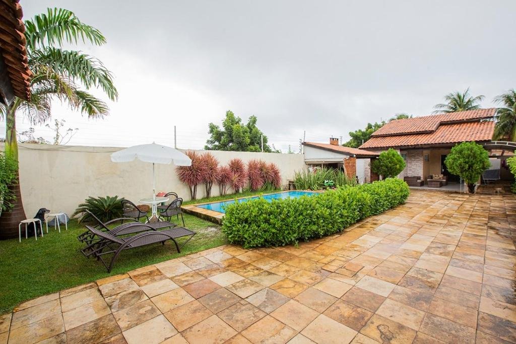 Quintal e piscina da casa Porto das Dunas Casa Temporada Estilo e Conforto. A piscina está do lado esquerdo, rodeada por arbustos e um gramado. Perto dela estão algumas espreguiçadeiras, uma mesas com cadeiras e um guarda-sol. Do lado direito há apenas o piso. Ao fundo está a casa.