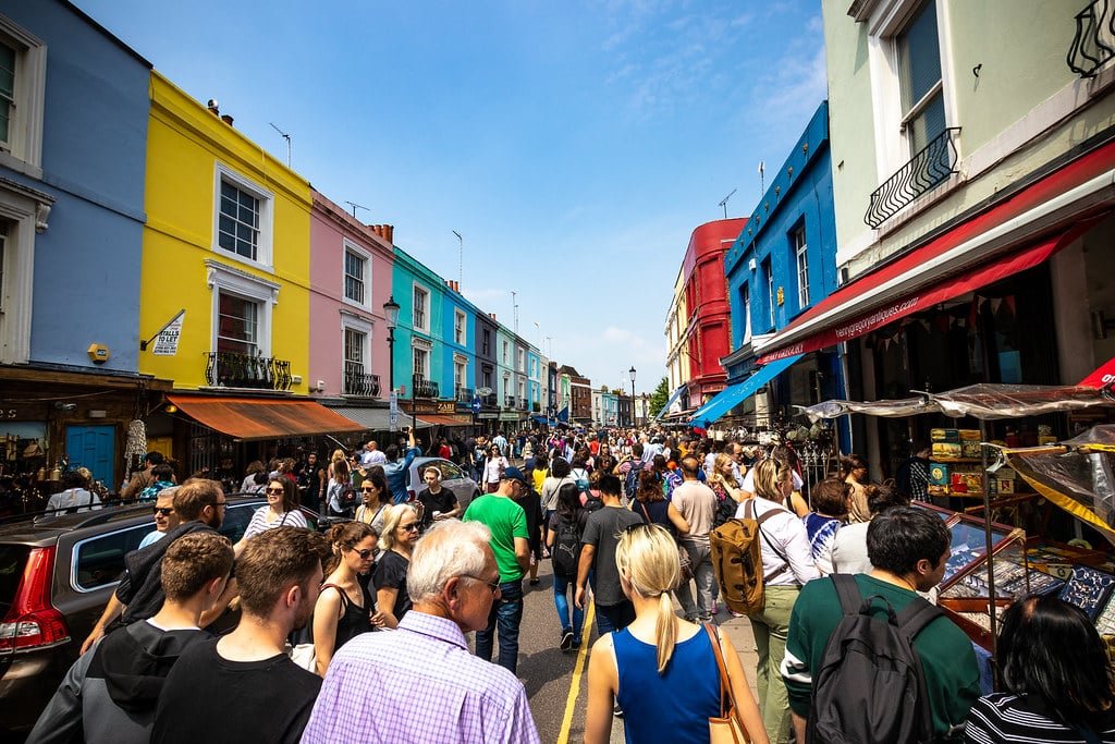 Portobello Road Market, um mercado de rua lotado com prédios coloridos, pessoas caminhando e várias barracas em um dia ensolarado.