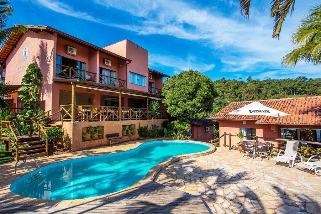 Parte da Pousada Altamira que mostra a piscina ao ar livre e do lado direito uma parte da pousada e alguma espreguiçadeiras e guarda-sol. Ao fundo da piscina tem outra construção de dois andares da pousada e em volta bastante árvores verdes durante o dia. Imagem ilustrando post Pousadas pet friendly no Litoral Norte de São Paulo.