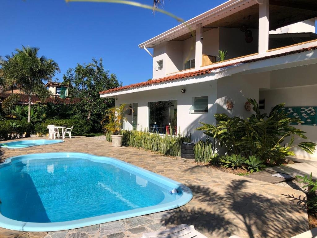 Parte da Pousada Baía das Conchas que mostra do lado esquerdo duas piscinas ao ar livre e do lado direito a construção de dois andares da pousada. Em volta tem algumas árvores e plantas verdes. Imagem ilustrando post Pousadas pet friendly no Litoral Norte de São Paulo.