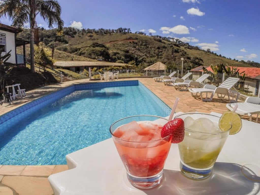 Foto da Pousada Campestre São Lourenço.  Na foto, há dois coquetéis em primeiro plano, um com uma morango e o outro com uma fatia de limão, ambos colocados em uma mesa branca; ao fundo, vemos uma piscina azul cercada por cadeiras brancas e guarda-sóis, com colinas verdes e um céu azul claro ao fundo.
