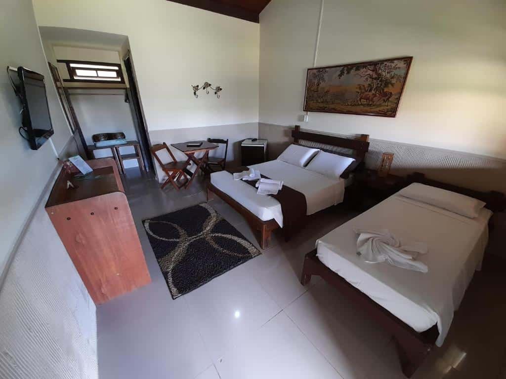 Foto da Pousada Campestre São Lourenço que está ilustrando o post de pousadas pet friendly em Minas Gerais. Na foto, há um quarto de hotel bem iluminado e organizado. À esquerda, há uma televisão pendurada na parede e um pequeno balcão de madeira abaixo dela. No centro do quarto, há duas camas de solteiro com lençóis brancos e travesseiros. Entre as camas, há uma pequena mesa com uma lâmpada. À direita das camas, há uma pintura pendurada na parede. No chão, entre a televisão e as camas, há um tapete preto e branco com um design geométrico. Ao fundo à esquerda, vemos parte de uma porta aberta que leva a outro cômodo.