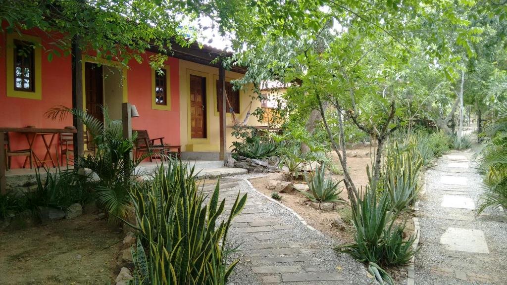 Área externa da Pousada Casa da Gente com ampla área verde, árvores, chão de pedrinhas e casas ao fundo onde ficam os quartos da pousada. Na varanda dessas pequenas casas ficam uma mesa com cadeiras ou uma rede.