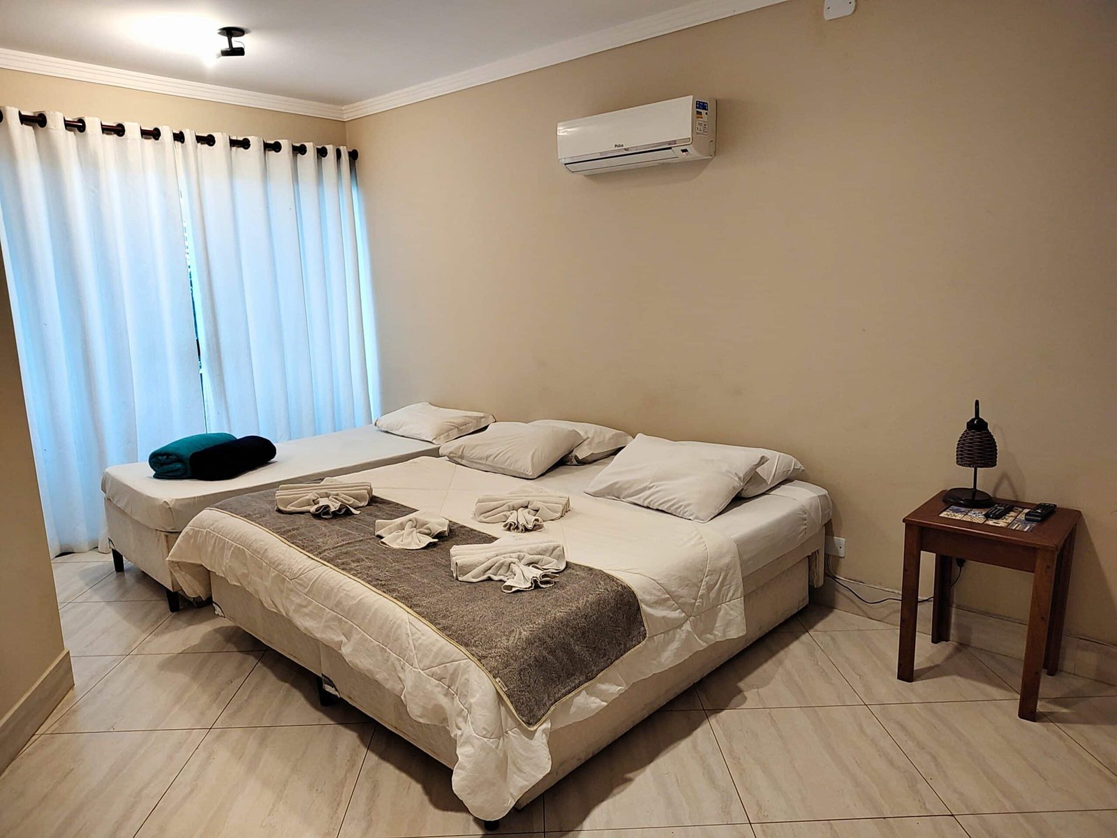 Quarto da Pousada Sol e Cia Tur com uma cama de casal e do lado uma cama de solteiro. Do lado da cama de casal tem uma mesa de cabeceira com um abajur e do lado da cama de solteiro tem uma cortina.