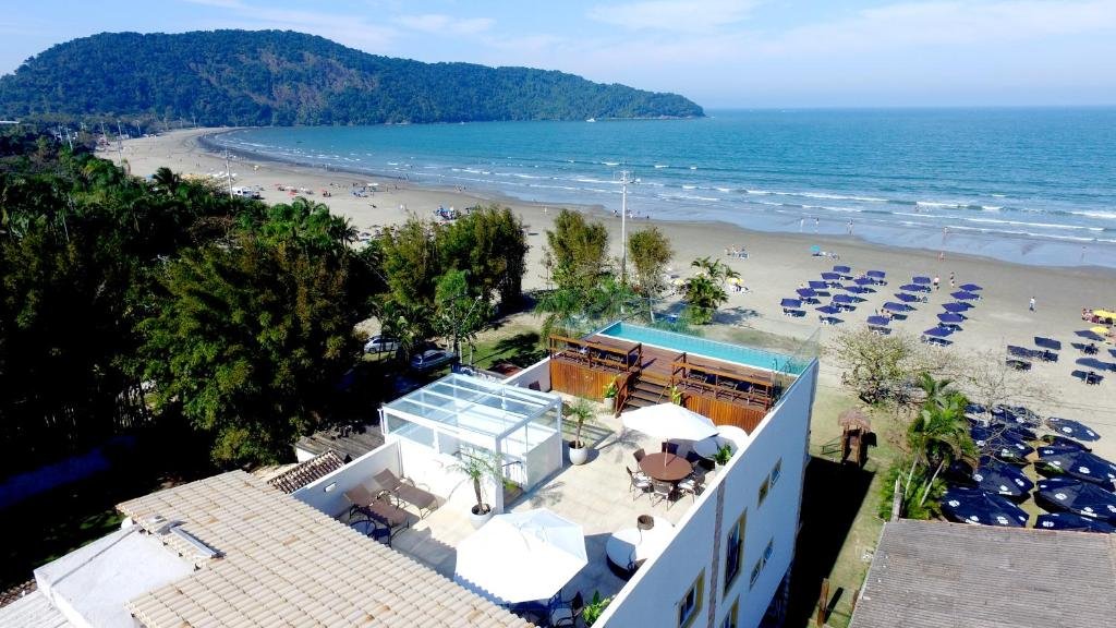 Foto aérea da Pousada Conca di Mare que mostra a construção da pousada bem de frente para a praia de faixa de areia extensa e grande mar com ondas. Já ao lado esquerdo da pousada tem bastante árvores verdes altas durante o dia. Imagem ilustrando post Pousadas pet friendly no Litoral Norte de São Paulo.