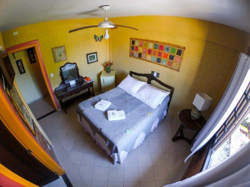 Quarto da Pousada das Laranjeiras. Uma cama no centro, do lado direito uma janela, uma mesa com abajur. Do lado esquerdo a porta do banheiro, a penteadeira e um frigobar. Foto para ilustrar post sobre pousadas pet friendly no Paraná.