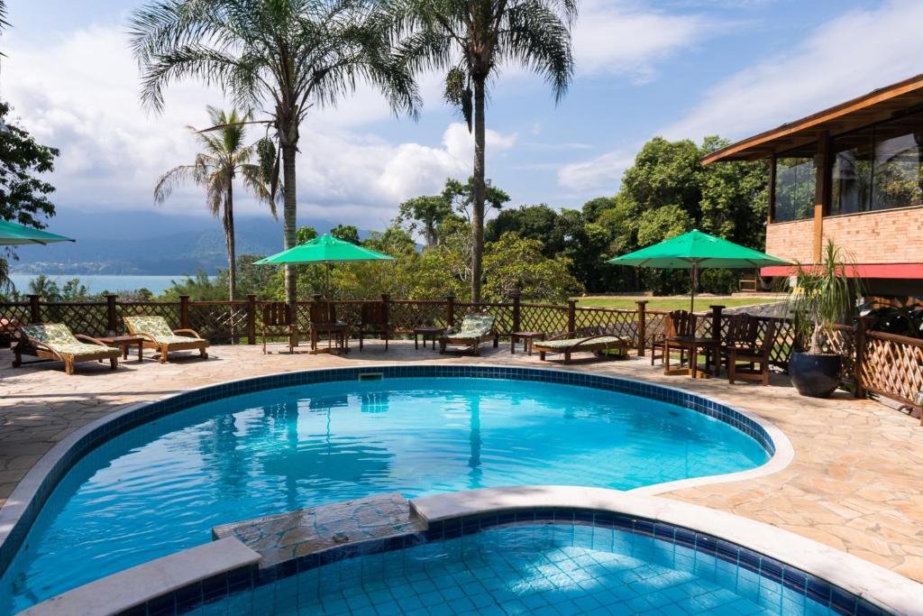 Área externa da Pousada Ilhote da Prainha. Pode-se ver ao centro duas piscinas, sendo uma maior e mais funda e a outra menor e mais rasa. Em volta da piscina há guarda-sóis, cadeiras, mesas e espreguiçadeiras, além de muitas árvores e natureza. Ao fundo é possível ver o mar.