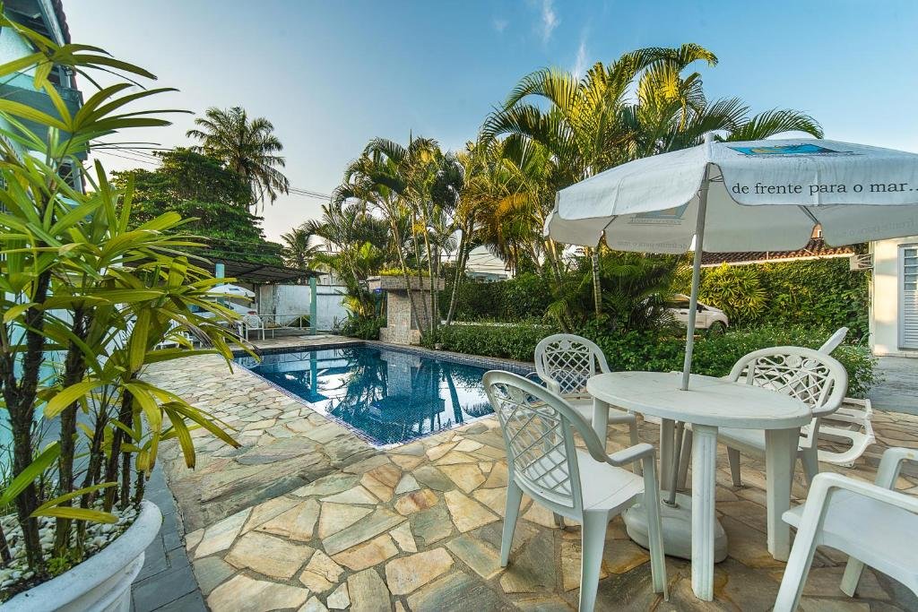 Parte da Pousada Porto do Itagua que mostra uma piscina ao ar livre e uma mesa com quatro cadeiras e um guarda-sol. Em volta tem bastante plantas e palmeiras. Imagem ilustrando post Pousadas pet friendly no Litoral Norte de São Paulo.