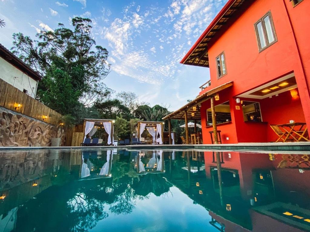 Parte da Pousada Porto Mare que mostra uma grande piscina ao ar livre e ao lado direito parte do bar da pousada e a construção de dois andares vermelhos da pousada. Ao fundo tem uma cadeiras, umas tendas e árvores. Imagem ilustrando post Pousadas pet friendly no Litoral Norte de São Paulo.