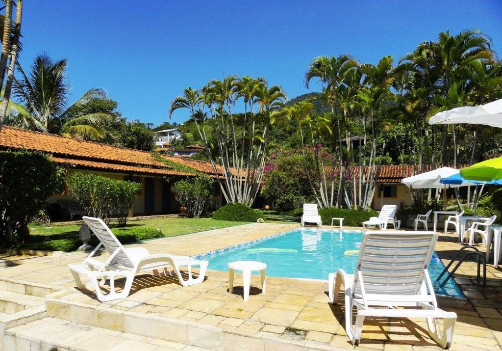 Parte da Pousada Praia do Curral que mostra uma piscina ao ar livre com algumas espreguiçadeiras e guarda-sóis em volta. Há também várias palmeiras e plantas e parte da construção da pousada. Imagem ilustrando post Pousadas pet friendly no Litoral Norte de São Paulo.