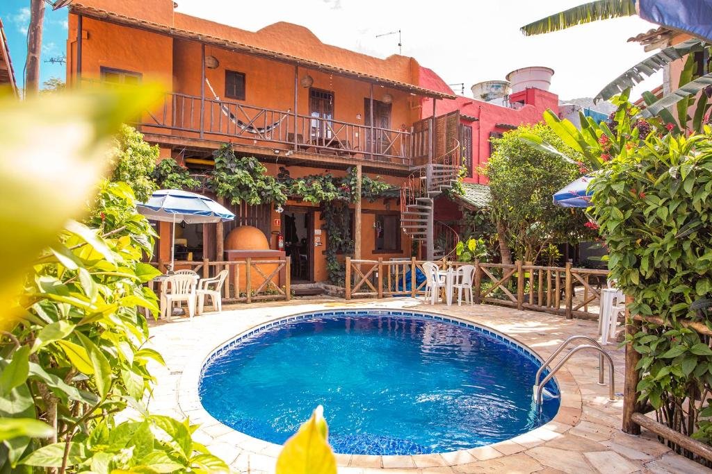 Área externa da Pousada Recanto da Villa. Ao centro há uma piscina circular, cercada por cadeiras, mesas e guarda-sóis. Em volta da área da piscina há plantas e arbustos. Ao fundo pode-se ver uma casa de dois andares com algumas das acomodações da pousada.