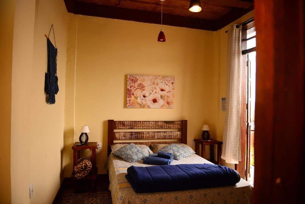 Foto da Pousada Residência Balestra que está ilustrando o post de pousadas pet friendly em Minas Gerais. A imagem mostra um quarto aconchegante com uma cama de casal, mesinhas de cabeceira, uma obra de arte na parede e uma porta que leva à luz natural, destacando-se pela sua simplicidade e conforto.