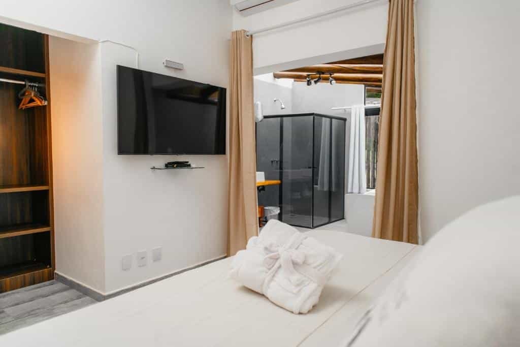 Foto da Pousada Röhsler que está ilustrando o post de pousadas pet friendly em Minas Gerais. A foto mostra um quarto moderno e bem iluminado, destacando-se pela organização e pelo design que integra a área de dormir e o banheiro. A cama está em um ângulo na qual vemos o quarto como se estivéssemos deitados nela. A TV está de frente, o banheiro à direita e um guarda-roupa está na esquerda.