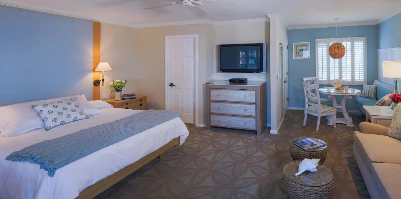 Quarto do Scripps Inn. Do lado direito um sofá, duas mesinhas de centro, no fundo uma mesa de jantar e janela. Do lado esquerdo uma cama de casal, duas cômodas e um abajur, uma TV e uma porta.