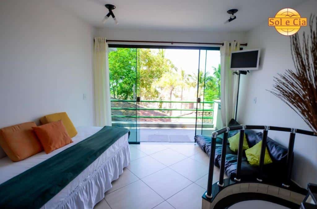 Sala da Pousada Sol e Cia Tur que mostra um sofá do lado esquerdo e do lado direito um sofá menor com uma tv na parede. Ao fundo tem uma porta grande de vidro com cortinas que dá acesso a varanda. Imagem ilustrando post Pousadas pet friendly no Litoral Norte de São Paulo.
