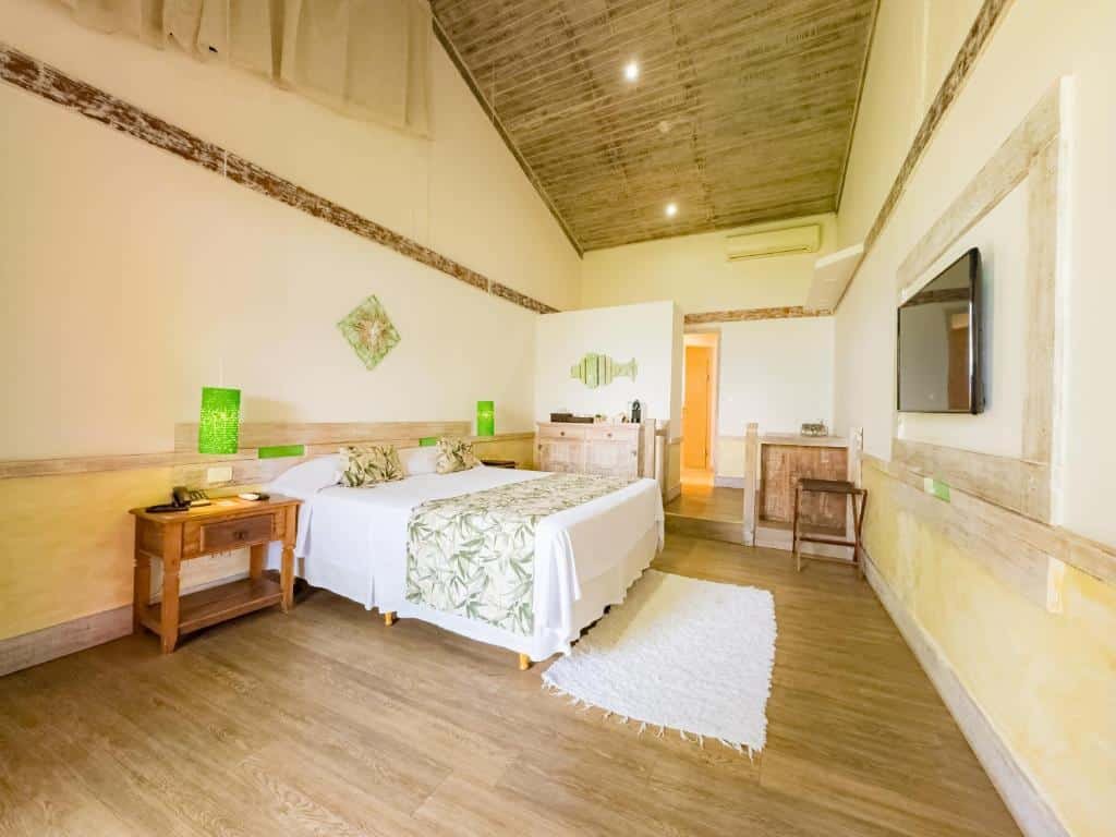 Pousada Vila das Velas. Ilustra o post sobre hotéis em Ilhabela. O quarto é espaçoso, com paredes claras e um teto de madeira escura.
Há uma cama grande no centro com lençóis brancos e uma colcha estampada de folhas verdes.
Duas luminárias verdes estão nas mesinhas de cabeceira de madeira em ambos os lados da cama.
Um tapete branco felpudo está parcialmente sob a cama.
Há uma cadeira de madeira e uma mesa à direita, com um espelho retangular acima dela na parede.
Uma obra de arte verde que parece ser feita de vidro ou cerâmica está pendurada na parede à esquerda da cama.
O piso é feito de tábuas largas de madeira clara, complementando as cores claras das paredes e do teto.