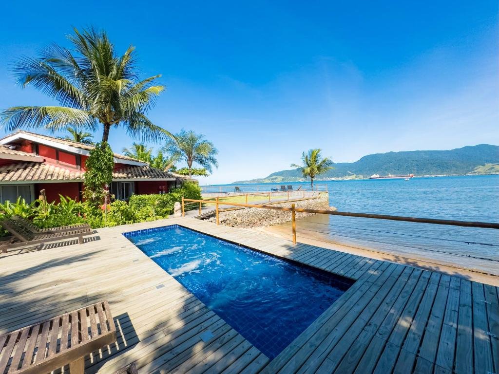 Pousada Vila das Velas. Há uma piscina retangular com água azul cristalina, cercada por um deck de madeira.
Vegetação: Uma palmeira alta e exuberante está próxima à piscina, adicionando um toque tropical ao cenário.
Edifício: O edifício visível tem um telhado vermelho e paredes brancas; parece ser parte de uma estrutura maior ou é uma cabana individual.
Cerca: Uma cerca de madeira delimita a área do deck, oferecendo segurança sem obstruir a vista espetacular do oceano e das montanhas ao fundo.
Céu: O céu está claro e azul, indicando um dia ensolarado e perfeito para relaxar ao ar livre.
Essa imagem transmite uma sensação de tranquilidade e férias à beira-mar.