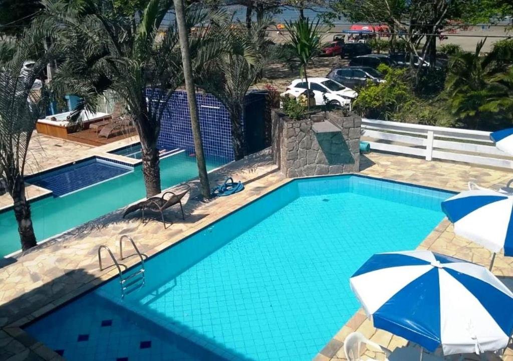 Parte da Pousada Vistazul que mostra duas piscinas ao ar livre com alguns guarda-sóis, espreguiçadeiras e palmeiras em volta. Esse espaço da pousada fica de frente para a rua do mar. Imagem ilustrando post Pousadas pet friendly no Litoral Norte de São Paulo.