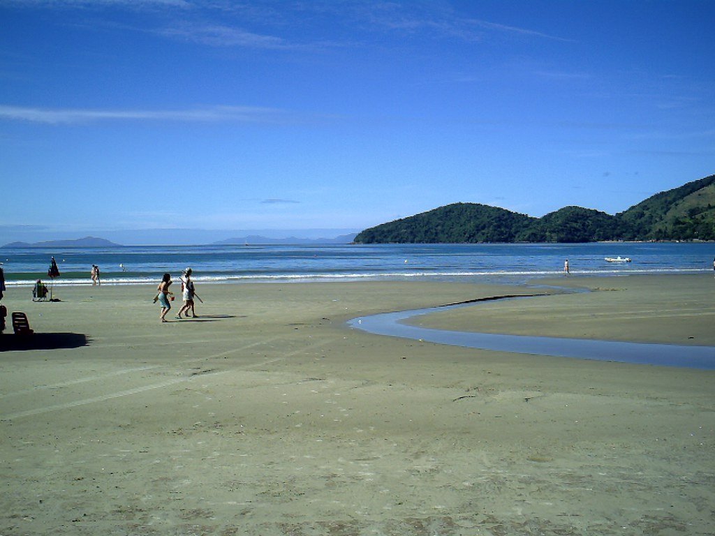 Praia da Enseada em Ubatuba. Pessoas caminhando, uma areia plana com um caminho de água do lado direito. Um mar calmo e com poucas ondas.