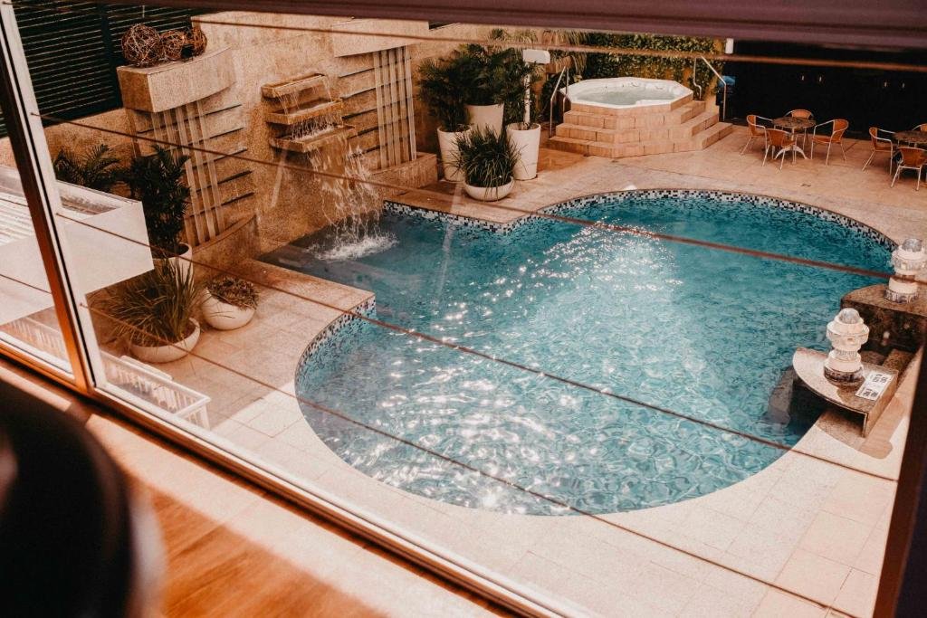 Piscina do Praiano Hotel em Fortaleza. Ela é redonda e possui uma cachoeria artificial na parede. Ao fundo há também uma banheira de hidromassagem. Ao redor existem algumas cadeiras e vasos de plantas.