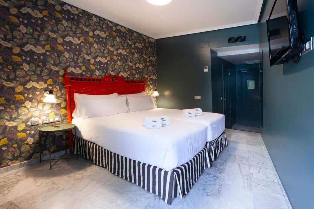Quarto do Petit Palace Puerta de Triana. Uma cama de casal com uma cômoda de cada lado e uma televisão de frente. Cabeceira vermelha e um papel de parede florido com fundo escuro.