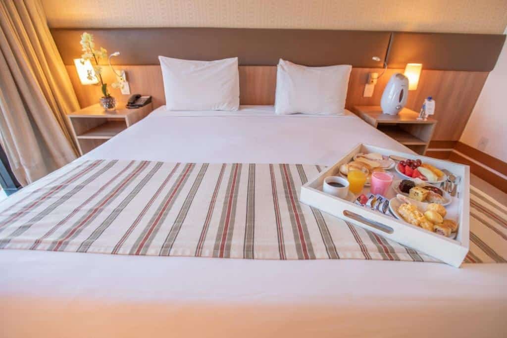 Quarto do Athos Bulcão Hplus Executive com uma cama de casal e um café da manhã servido dentro de uma bandeja, além de duas mesas de cabeceira com luminárias, para representar hotéis perto do consulado americano em Brasília