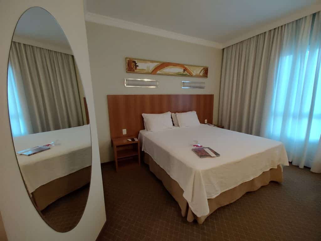 suíte do Bourbon Joinville Convention Hotel com uma cama de casal à direita da imagem, no centro do quarto. À esquerda é possivel ver uma parede com um espelho oval na vertical