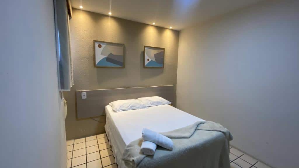 Quarto do Center Plaza Hotel. A cama está centralizada com um cobertor e duas toalhas em cima, atrás da cama fica uma cabeceira de madeira e dois quadros decorativos em cima. Ao lado esquerdo é possível ver uma cortina que vai até metade da parede. Imagem para ilustrar o post hotéis em Caruaru.