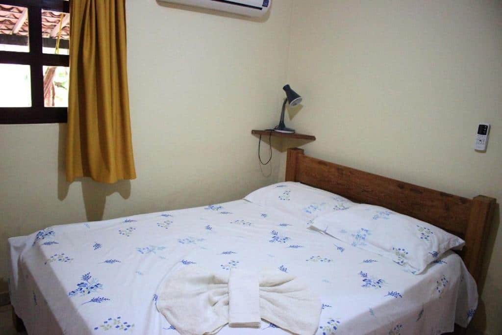 Quarto da Pousada Casa da Gente. Na foto é possível ver uma cama de casal centralizada, uma prateleira pequena com um abajur no lado esquerdo da cama. também é possível ver uma parte da janela no lado esquerdo. Imagem para ilustrar o post hotéis em Caruaru.