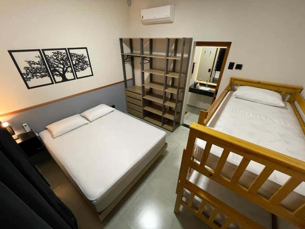 Quarto do Cantinho Estrelado com uma cama de casal com dois travesseiros, um beliche de madeira e do lado um guarda-roupa aberto com um ar-condicionado em cima. Do lado do guarda-roupa tem uma porta que dá acesso ao banheiro do quarto.
