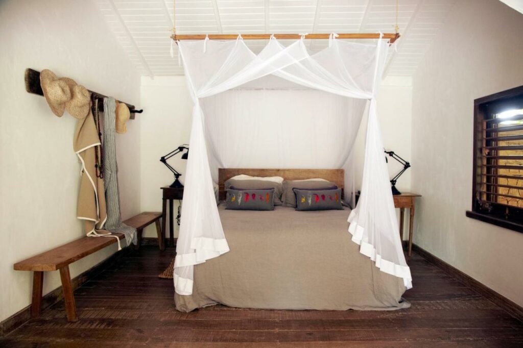 Quarto da Casa Angatu com cama de casal no centro do quarto, uma cômoda em cada lado da cama com luminária e do lado esquerdo da cama um banco de madeira.