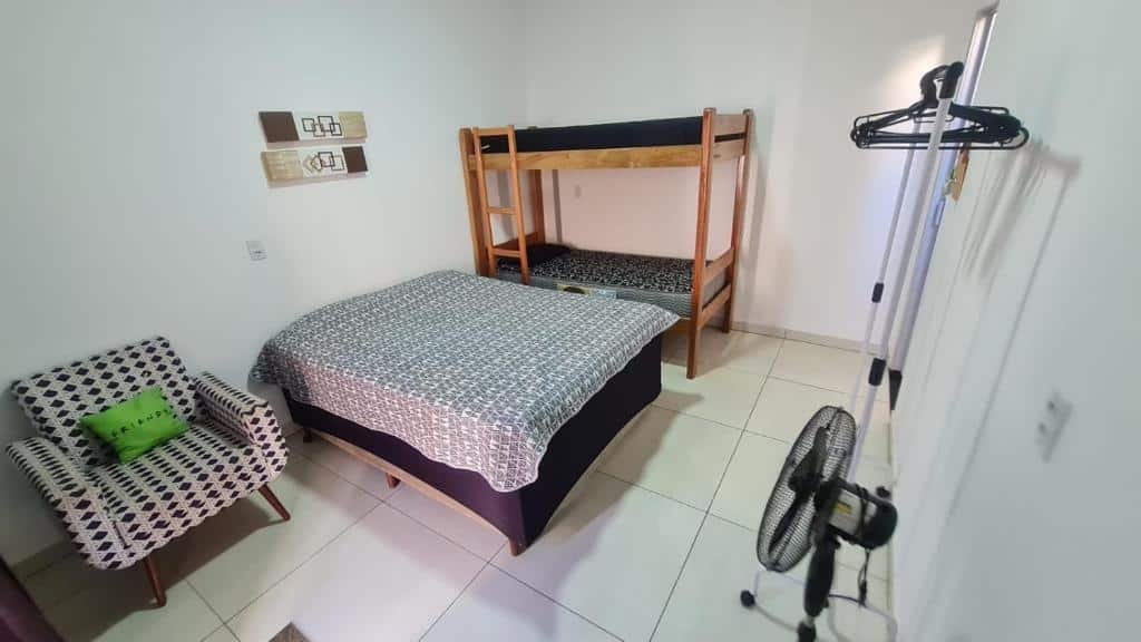 Quarto do Casa c/ quartos separados + churrasqueira ampla com uma cama de casal ao centro do quarto, do lado esquerdo uma poltrona e do lado direito um beliche de madeira. No quarto tem uma arara de roupas e um ventilador no chão.