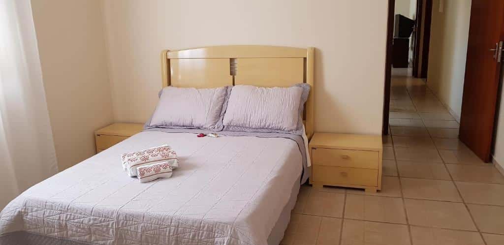 Quarto da Casa da Cachoeira" com uma cama de casal com dois travesseiros, uma colcha e duas toalhas em cima. De cada lado da cama tem uma mesinha e do lado direito da foto tem uma porta aberta que dá acesso ao corredor da casa.