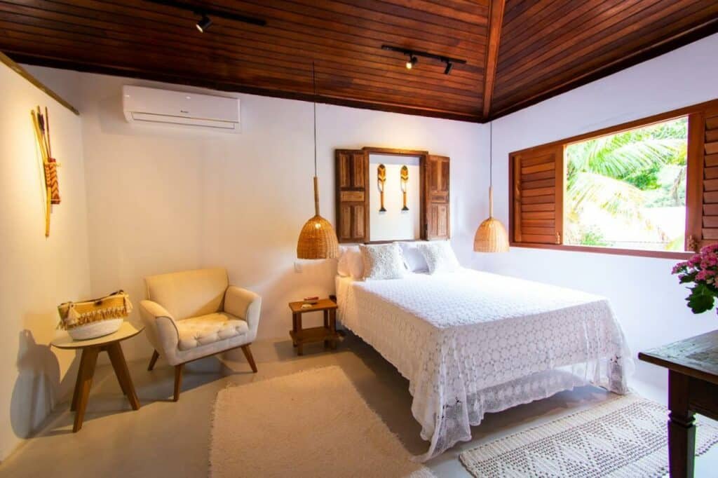 Quarto da Casa João Viera Trancoso com cama de casal do lado esquerdo da imagem com uma cômoda do lado direito da cama, onde também se encontra uma poltrona e uma mesa. Representa airbnb em Trancoso.