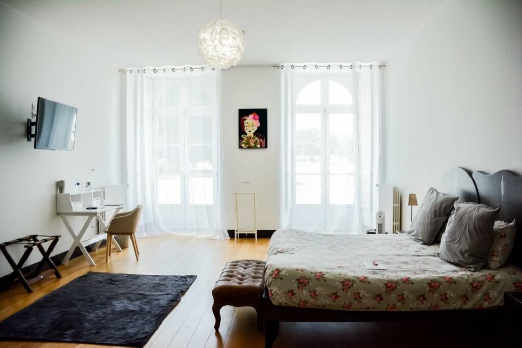 Quarto do Chateau de Briançon com cama de casal do lado direito da imagem, no pé da cama um banco estofado em frente a cama uma TV presa na parede e do lado esquerdo da TV uma mesa de trabalho com cadeira. Representa hotéis no Vale do Loire.