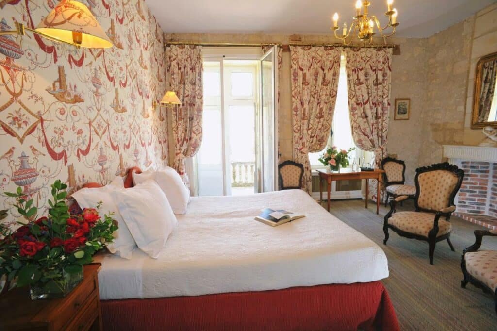 Quarto do Château De La Bourdaisière, com cama de casal do lado esquerdo da imagem com uma cômoda do lado direito da cama com um vaso de flores e na frente da cama duas cadeiras. Representa hotéis no Vale do Loire.