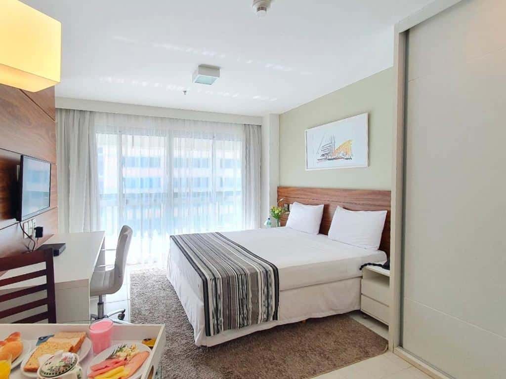 Quarto no Fusion Hplus Express + com uma sacada com cortinas, uma cama de casal, um tapete, na parede de frente com a cama há uma televisão e uma mesa de escritório com cadeira giratória, além de uma outra mesa para refeições com dois lugares, para representar hotéis perto do consulado americano em Brasília