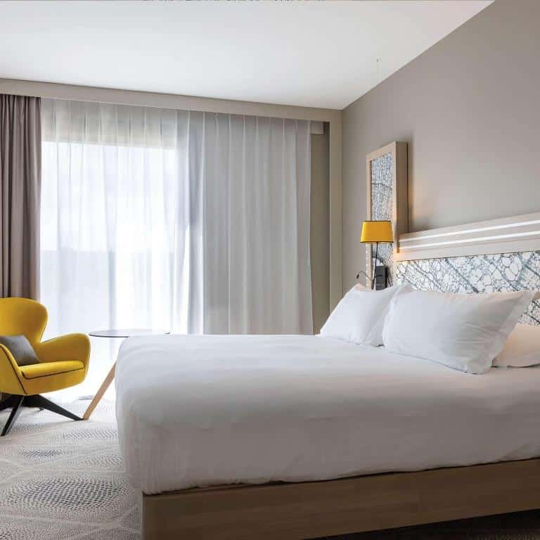 Quarto do Hilton Garden Inn Tours Centre, France com cama de casal do lado direito da imagem e em frente a cama uma cadeira. Representa hotéis no Vale do Loire.