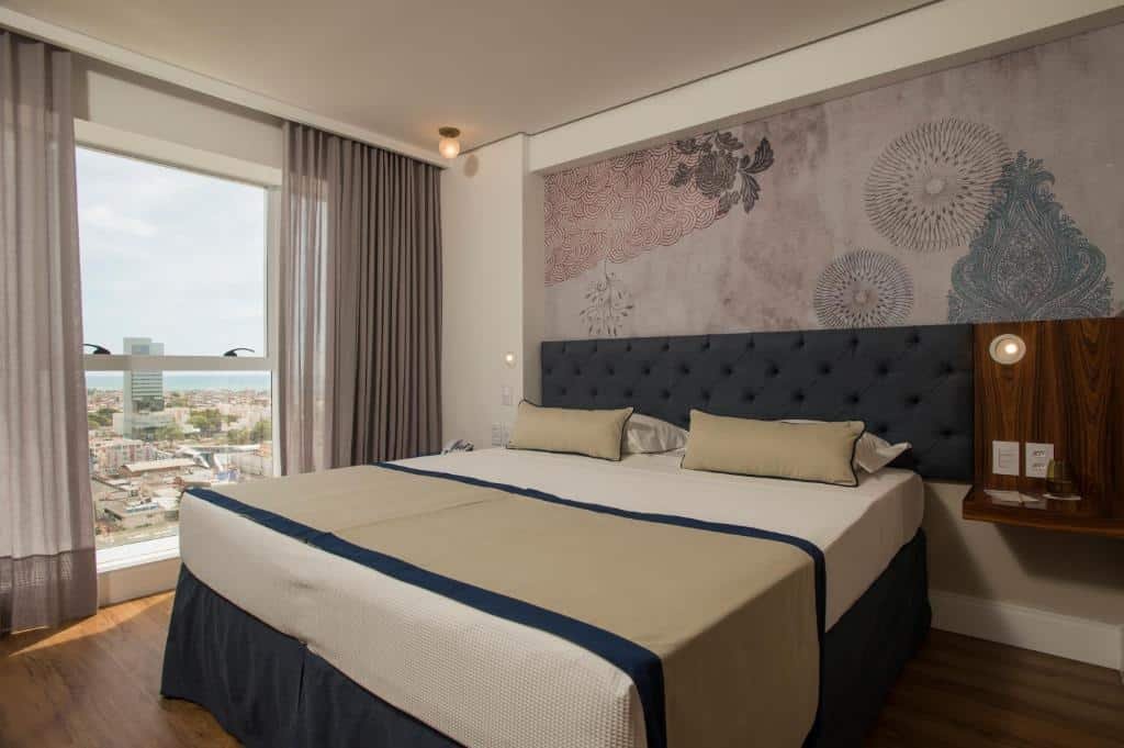 Uma cama com uma cômoda do lado. Na parede do lado esquerdo uma janela do tamanho da parede com vista para a cidade.