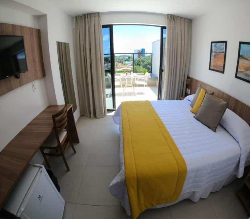 Uma cama de casal no lado direito. Um frigobar, uma mesa de escritório e uma televisão no lado esquerdo. Uma sacada no quarto. Foto para ilustrar post sobre hotéis perto do Consulado Americano em Recife.