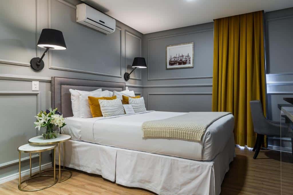 Uma cama de casal com um abajur de cada lado e um ar-condicionado em cima. Na frente, uma mesa de escritóri.