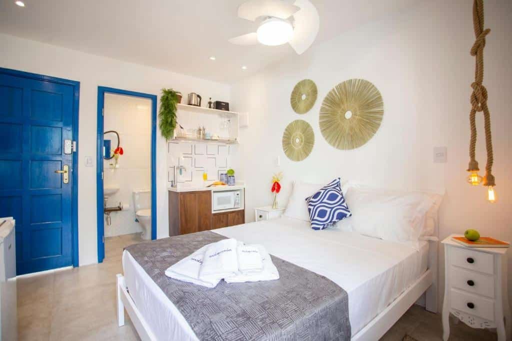 Uma cama de casal com uma cômoda do lado. Um armário do lado com microondas e utensílios de cozinha. Ao lado do armário a porta do banheiro.