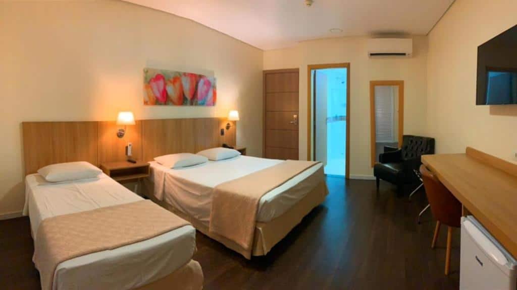 Uma cama de casal e uma cama de solteiro no lado esquerdo. No lado direito uma mesa de escritório, um frigobar, uma televisão e uma poltrona.