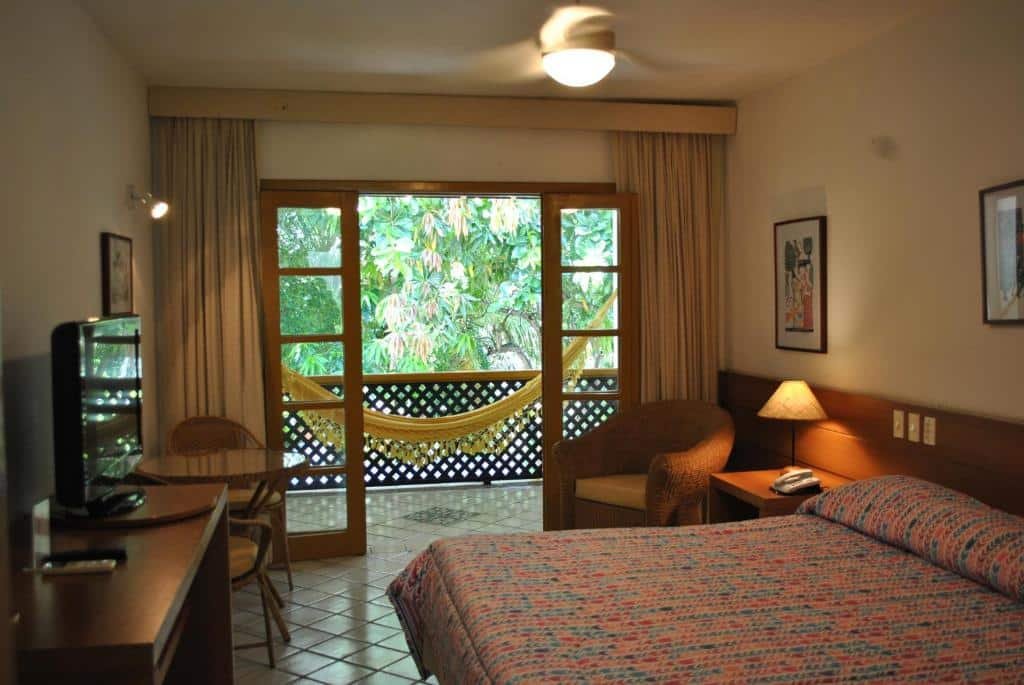 Uma cama de casal no canto direito com uma cômoda e uma poltrona. No canto esquerdo uma televisão e uma mesa com cadeira. Atrás uma sacada com uma rede.